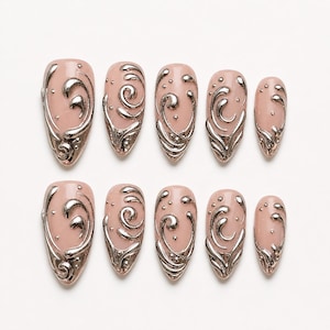 May include: A set of ten press-on nails featuring a light pink base colour with raised, metallic silver swirling patterns and small silver dots. The nails have an almond shape and are arranged in two rows of five, decreasing in size from left to right.