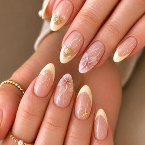 May include: Almond-shaped nails featuring a French manicure with pale yellow tips. The design includes clear base coats adorned with gold starfish charms, small white pearls, a gold seashell accent, and delicate hand-painted white floral art.
