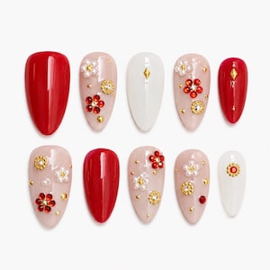 May include: A set of ten almond-shaped press-on nails featuring a mix of solid red and white glossy finishes, accented with small white pearl flowers, red crystal floral embellishments, gold metallic studs, and tiny clear rhinestones.