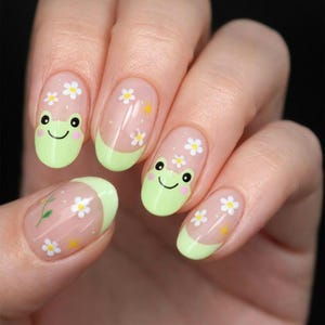 May include: Nail art featuring a clear base coat with light green French tips. Two nails display a cartoon frog face with black eyes and a smiling mouth. All nails are decorated with small white daisy flowers and tiny yellow star accents.