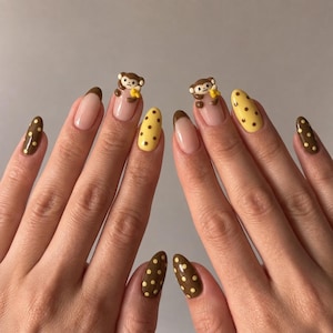 May include: Almond-shaped nails featuring a mix of designs including brown tips on a nude base, solid yellow nails with brown polka dots, and brown nails with yellow polka dots. Two nails feature a three-dimensional brown monkey holding a yellow banana.