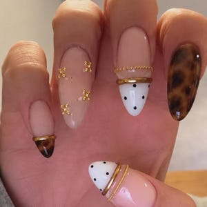 May include: Almond-shaped nails featuring a mix of designs including a nude base with small gold cross accents, a white tip with black polka dots and gold bands, and a solid brown tortoiseshell pattern on a full nail and a single nail tip.