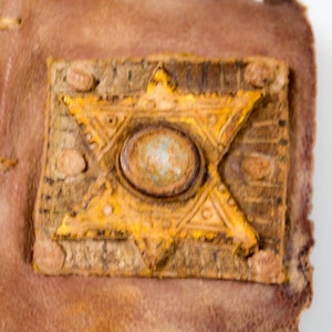 Puede incluir: Primer plano de una cubierta de libro de cuero marr&oacute;n con un dise&ntilde;o de estrella de David amarilla y una piedra redonda azul verdosa en el centro.