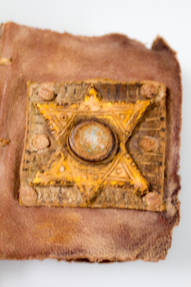 Puede incluir: Primer plano de una cubierta de libro de cuero marr&oacute;n con un dise&ntilde;o de estrella de David amarilla y una piedra redonda azul verdosa en el centro.