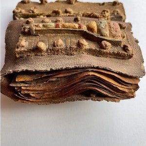 Puede incluir: Un primer plano de un antiguo libro encuadernado en cuero con una cubierta decorativa. La cubierta es de cuero marr&oacute;n y tiene un dise&ntilde;o en relieve de una figura estilizada. El libro est&aacute; cerrado y las p&aacute;ginas son visibles a trav&eacute;s de los bordes abiertos.