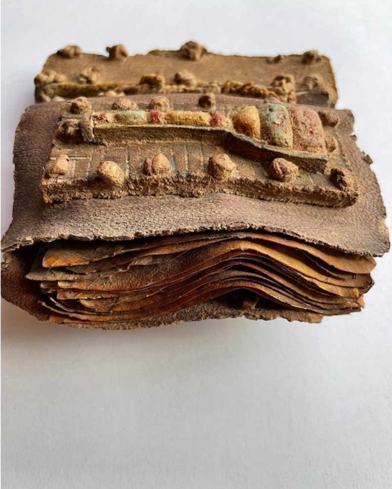 Puede incluir: Un primer plano de un antiguo libro encuadernado en cuero con una cubierta decorativa. La cubierta es de cuero marr&oacute;n y tiene un dise&ntilde;o en relieve de una figura estilizada. El libro est&aacute; cerrado y las p&aacute;ginas son visibles a trav&eacute;s de los bordes abiertos.