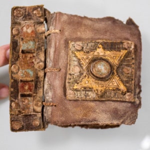 Puede incluir: Un peque&ntilde;o libro encuadernado en cuero marr&oacute;n con un dise&ntilde;o de estrella de seis puntas en la portada. La estrella es de metal y tiene un centro circular met&aacute;lico.