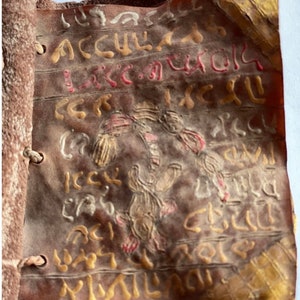 Puede incluir: Un primer plano de un libro encuadernado en cuero con letras e ilustraciones pintadas en oro y rojo. El libro es viejo y est&aacute; desgastado, faltan algunas p&aacute;ginas. El texto est&aacute; en un idioma antiguo.