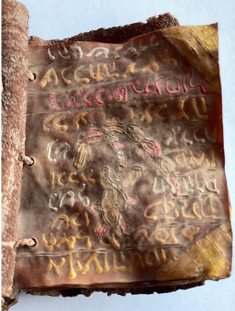 Puede incluir: Un primer plano de un libro encuadernado en cuero con letras e ilustraciones pintadas en oro y rojo. El libro es viejo y est&aacute; desgastado, faltan algunas p&aacute;ginas. El texto est&aacute; en un idioma antiguo.