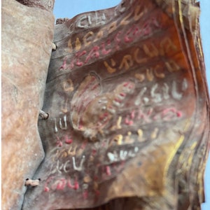 Puede incluir: Un primer plano de un viejo libro encuadernado en cuero con p&aacute;ginas llenas de texto manuscrito descolorido en tinta dorada, roja y blanca. El libro est&aacute; desgastado y tiene un aspecto r&uacute;stico.