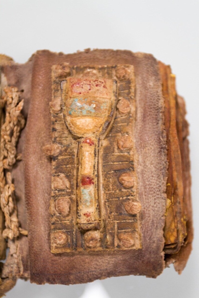 Puede incluir: Primer plano de un libro encuadernado en cuero con un cierre decorativo. El cierre es de metal y tiene un dise&ntilde;o de un objeto estilizado con pintura roja, azul y blanca.