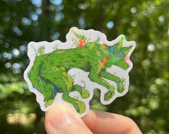 May include: A sticker featuring a green wolf-like creature covered in foliage. The sticker has a white border and is held in a hand against a blurred green background. The wolf has colorful accents.