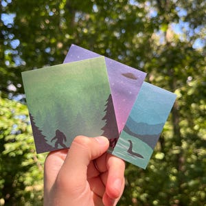 Puede incluir: Una mano sostiene cuatro tarjetas cuadradas con ilustraciones de críptidos. Una tarjeta muestra una silueta de Bigfoot en un bosque, otra un OVNI y una tercera el monstruo del lago Ness. Las tarjetas tienen un degradado de color.