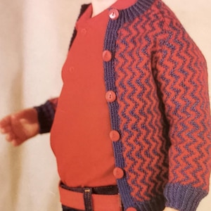 以下が含まれることがあります： ボタン付きの赤と青の模様のカーディガンを着た幼児。赤いシャツを着て、青いジーンズと赤いベルトをしています。