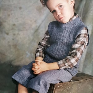 May include: A young person in a studio portrait, wearing a blue knit vest over a plaid shirt, blue pants, and gray socks. The person is seated on a wooden box, with hands clasped. The background is a mottled gray.