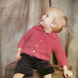 May include: A young child wearing a pink knitted cardigan with buttons and black shorts sits on a wooden crate. The child is looking up and smiling.