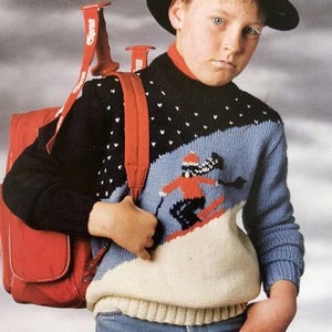 Può includere: Un giovane che indossa un cappello nero e un maglione con motivo blu, bianco e rosso. Porta uno zaino rosso con una tracolla bianca su cui è scritto "Simon".
