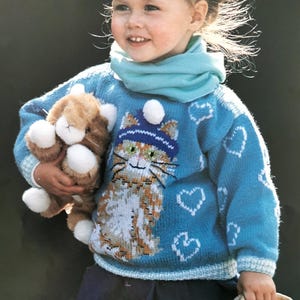 May include: A young girl wearing a blue knitted sweater with a cat design and white hearts. She is holding a brown and white stuffed animal and a wicker basket.