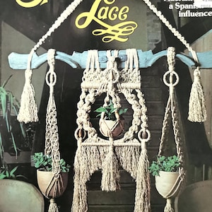 May include: A macrame plant hanger with a Spanish influence, inspired by "Las Calles". The hanger is made of natural fiber rope and has a wooden top bar. The hanger is decorated with tassels and has three plant pots.