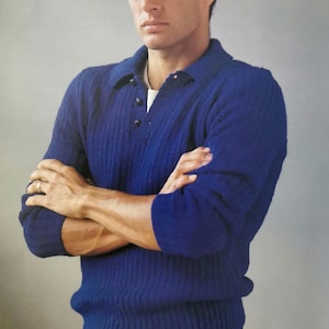 May include: A man wearing a blue sweater with a button-up collar and black pants with the text "NYAC" and a winged figure on the back.