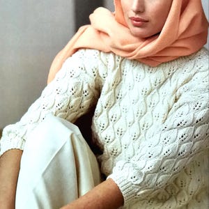 May include: A woman wearing a white knitted sweater with a delicate openwork pattern. She is wearing a peach-colored scarf around her neck and white pants.
