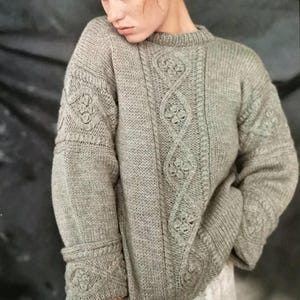 Puede incluir: Un suéter de punto gris con un diseño de punto de cable. El suéter tiene un cuello redondo y mangas largas.