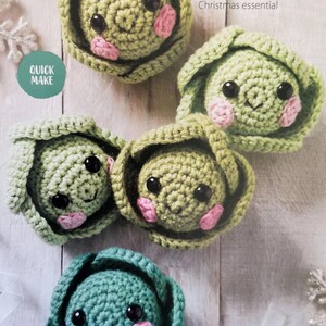 May include: Five crocheted amigurumi Brussels sprout ornaments with smiling faces. Each sprout is green with pink cheeks and black eyes. The ornaments are arranged on a light wood background.