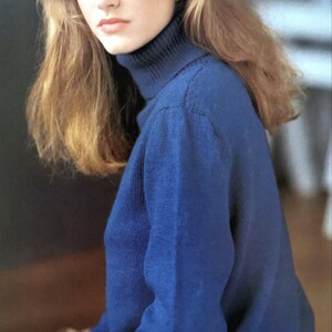 May include: A woman with long blonde hair is wearing a blue turtleneck sweater. The sweater is knitted and has a ribbed texture.