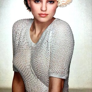 May include: A woman wearing a light beige, open-weave knit top with short sleeves and a matching tie at the waist. A large cream-colored flower adorns her hair. The image has a soft, vintage aesthetic.