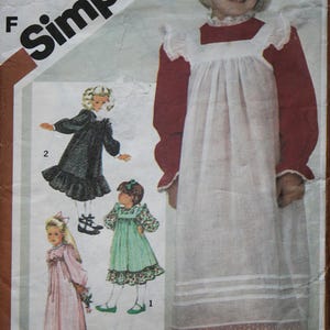 May include: A vintage Simplicity sewing pattern for a child's dress, size 5. The pattern includes illustrations of different dress designs, such as a long white dress with red sleeves and trim. The package features the Simplicity logo.