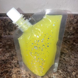 May include: A clear plastic pouch filled with a yellow liquid and silver glitter. The pouch has a white plastic spout.