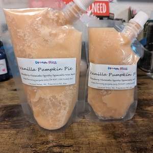 May include: Two clear pouches filled with a light orange, textured substance. Each pouch has a white label with the text "Dream Bliss Vanilla Pumpkin Pie." The pouches have white spouts. The text "Blueberry Cheesecake Squishy Squeezable Wax Melt" is also visible.