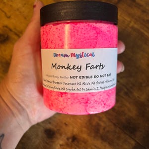 May include: A jar of pink whipped body butter with the text "Monkey Farts" and "Dream Mystical" on the label. The jar has a black lid and contains a pink, speckled substance. The label also lists ingredients.