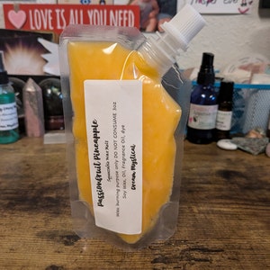 May include: A yellow squeezable pouch of passionfruit pineapple wax melt. The pouch is labeled with the product name, "Passionfruit Pineapple", and the instructions "Squeezable Wax Melt, Wax burning purpose only DO NOT CONSUME 3 oz, Soy Wax, Oil, Fragrance Oil, dye, Dream Mystical."