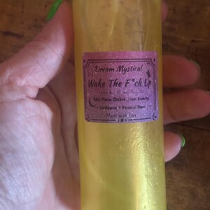 Puede incluir: Una botella de spray llena de líquido amarillo con una tapa blanca. La etiqueta dice "Dream Mystical Wake The F*ck Up" con texto sobre chakras y confianza. La botella se sostiene en una mano.