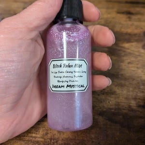 May include: A clear glass bottle with a pink liquid and glitter inside. The bottle has a black spray top and a white label that reads "Bitch Relax Mist" and "DREAM MYSTICAL".