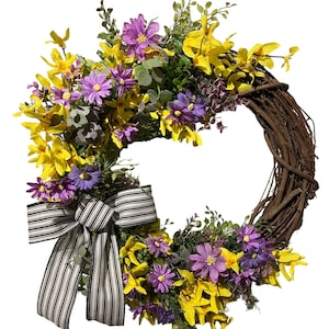 May include: A decorative wreath featuring a woven grapevine base adorned with yellow and purple flowers, green foliage, and a black and white striped bow. A spring or summer themed home decor item.