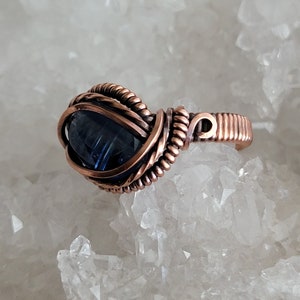 May include: A copper wire wrapped ring with a dark blue gemstone. The ring is set on a bed of white crystals.