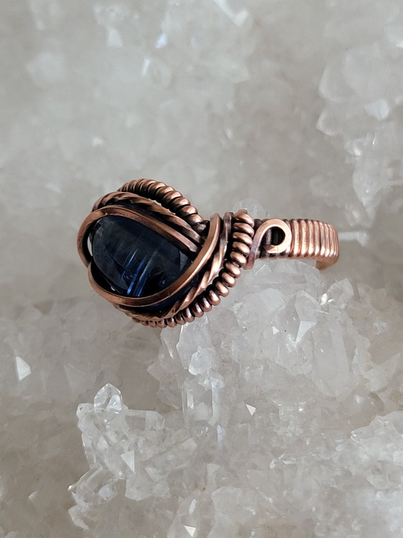 May include: A copper wire wrapped ring with a dark blue gemstone. The ring is set on a bed of white crystals.
