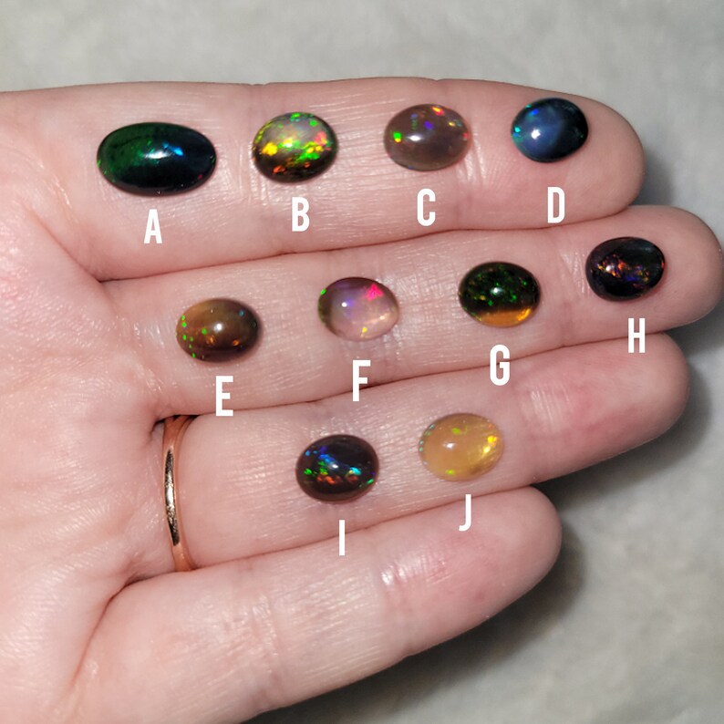 May include: Ten oval-shaped opals in various colors, including black, green, brown, and gold, are displayed on a hand. The opals are labeled with letters A through J.
