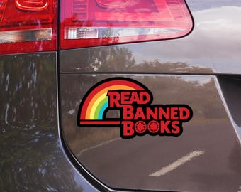 Puede incluir: Adhesivo negro con el texto "READ BANNED BOOKS" en rojo, con un diseño de arcoíris. La pegatina está adherida a la parte trasera de un coche gris oscuro, cerca del piloto trasero.