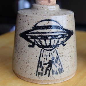 May include: A ceramic bottle with a cork stopper, featuring a black illustration of a UFO beaming up a person. The bottle has a speckled, tan finish.