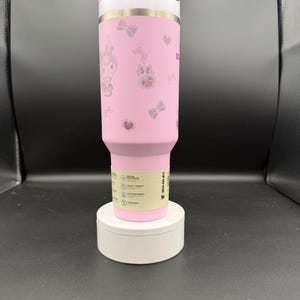 Puede incluir: Un vaso Stanley rosa con tapa blanca y pajita blanca. El vaso est&aacute; decorado con personajes de dibujos animados, lazos y corazones. El vaso est&aacute; sobre un pedestal blanco.