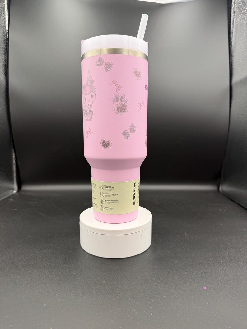 Puede incluir: Un vaso Stanley rosa con tapa blanca y pajita blanca. El vaso est&aacute; decorado con personajes de dibujos animados, lazos y corazones. El vaso est&aacute; sobre un pedestal blanco.