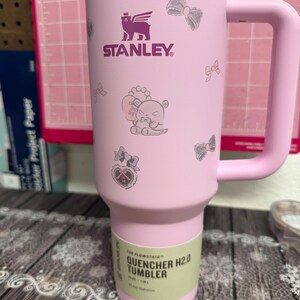 Puede incluir: Vaso Stanley Quencher H2.0 rosa con asa, con el logotipo morado de Stanley y lazos decorativos e ilustraci&oacute;n de oso. El vaso tiene una capacidad de 1,18L.