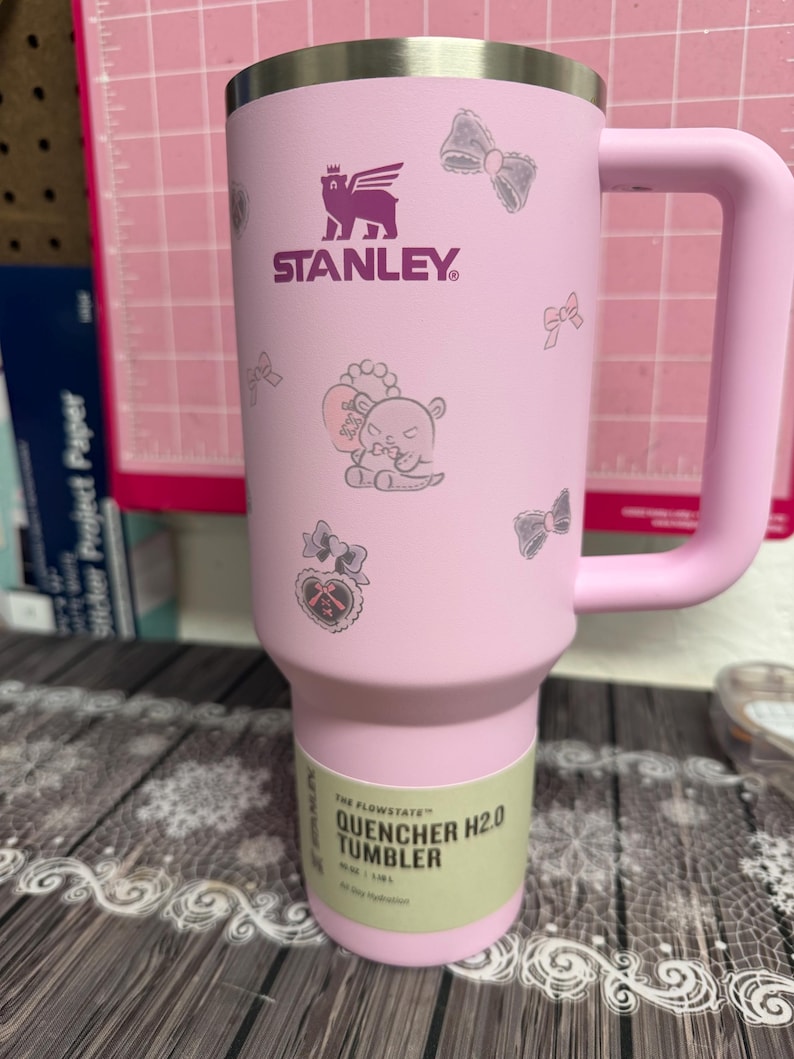 Puede incluir: Vaso Stanley Quencher H2.0 rosa con asa, con el logotipo morado de Stanley y lazos decorativos e ilustraci&oacute;n de oso. El vaso tiene una capacidad de 1,18L.