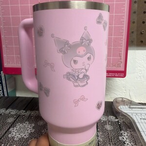 Puede incluir: Un vaso rosa de acero inoxidable con asa, decorado con un personaje de dibujos animados y motivos de lazo. El dise&ntilde;o incluye un personaje con calavera y lazo. El vaso tiene un borde plateado.
