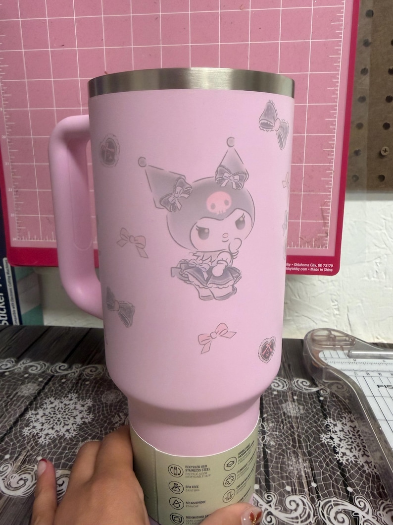 Puede incluir: Un vaso rosa de acero inoxidable con asa, decorado con un personaje de dibujos animados y motivos de lazo. El dise&ntilde;o incluye un personaje con calavera y lazo. El vaso tiene un borde plateado.