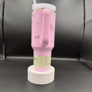 Puede incluir: Vaso t&eacute;rmico rosa con tapa y asa blancas. El vaso presenta un dise&ntilde;o de lazo y personaje. Incluye una pajita blanca. El vaso est&aacute; sobre una plataforma blanca.