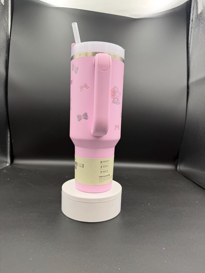 Puede incluir: Vaso t&eacute;rmico rosa con tapa y asa blancas. El vaso presenta un dise&ntilde;o de lazo y personaje. Incluye una pajita blanca. El vaso est&aacute; sobre una plataforma blanca.